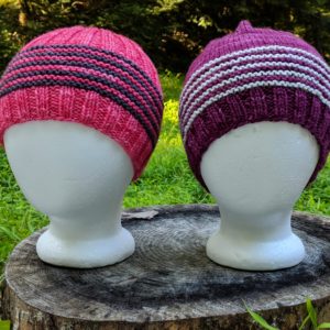 Two knit hats on styrofoam heads, facing forward, one a fitted beanie in pink with black stripes, the other a purple slouchy beanie with gray stripes.