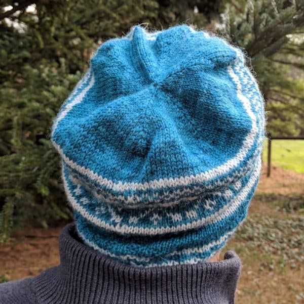 Back view showing crown decreases of a peacock blue slouchy beanie withlight gray geometric colorwork motifs.