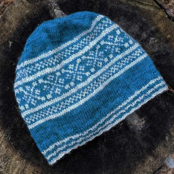 Flat view of a peacock blue slouchy beanie withlight gray geometric colorwork motifs.