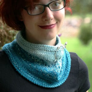 A gently smiling young woman with short red hair, wearing red lipstick, black framed glasses and a black shirt models a blue two-toned knitted cowl that features a speckled colorwork motif. It is folded over to be closer to her neck and is secured with a silver shawl pin.