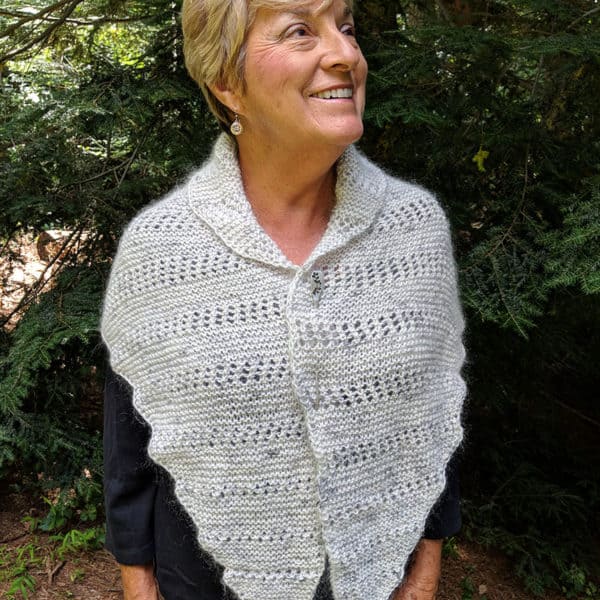 Smiling woman with short blonde hair modeling an asymmetrical, triangular light gray wrap featuring a lace motif. She is wearing it draped over her shoulders with a shawl collar effect and pinned with a silver shawl pin.